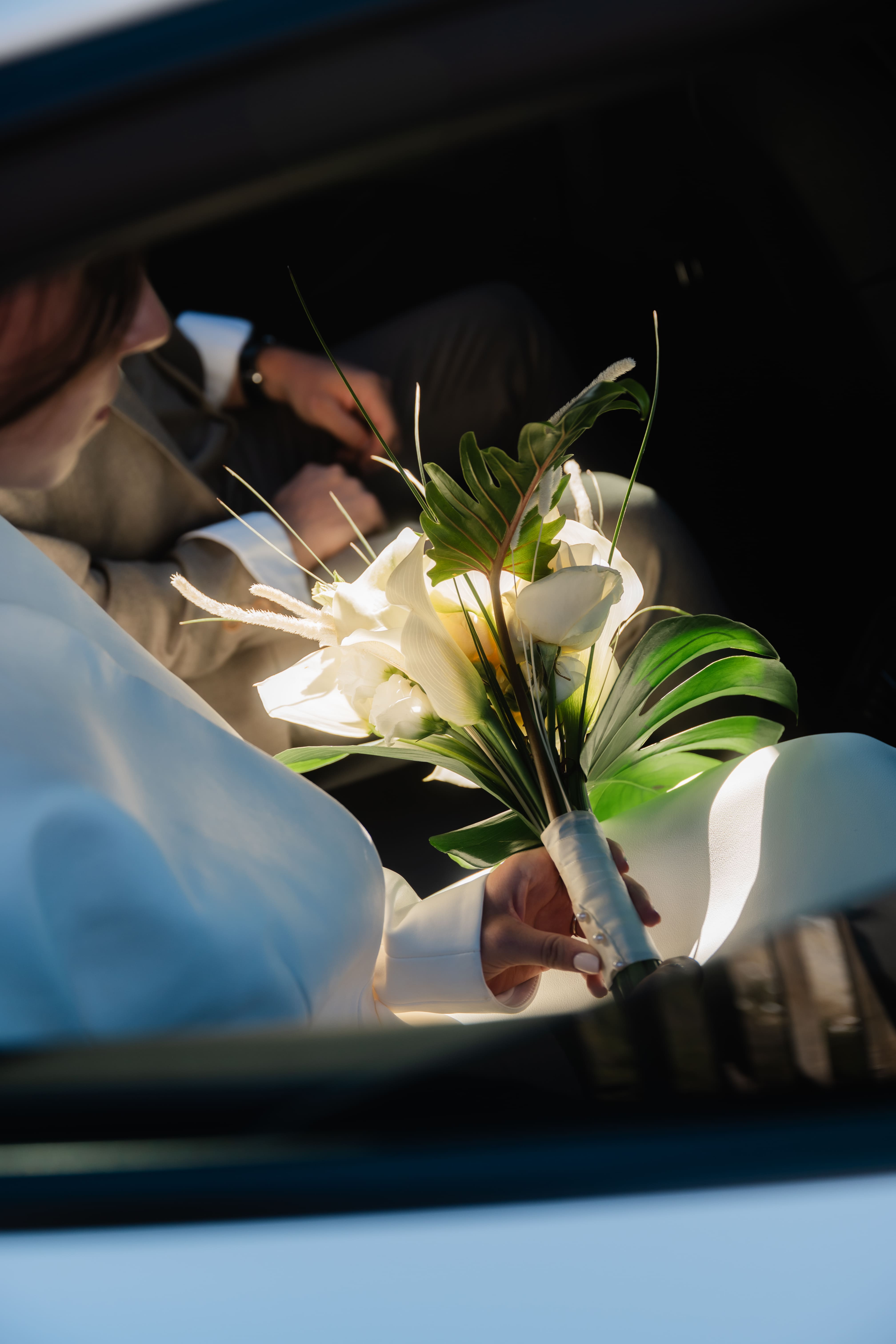 Bouquet de mariée blanc à feuilles de monstera tenu à travers la fenêtre d'une voiture, contre-jour.